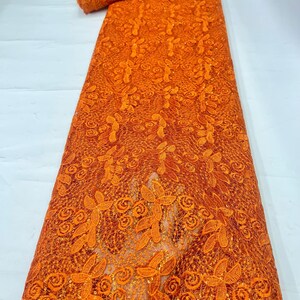 May include: A vibrant orange lace fabric featuring an intricate floral and leaf pattern. The textile is embellished with small, shimmering sequins throughout the design, creating a textured surface with a repeating botanical motif across the material.