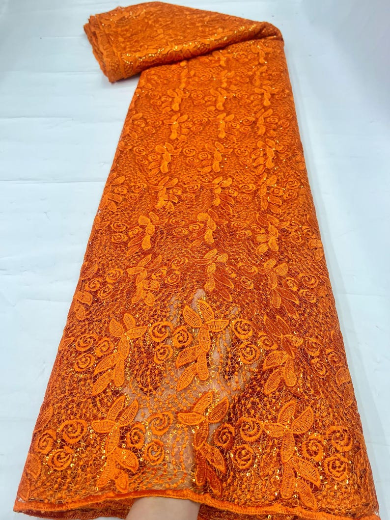 May include: A vibrant orange lace fabric featuring an intricate floral and leaf pattern. The textile is embellished with small, shimmering sequins throughout the design, creating a textured surface with a repeating botanical motif across the material.