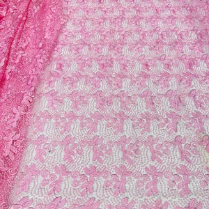 May include: A vibrant pink lace fabric featuring an intricate embroidered pattern of swirling floral motifs and leaves. The delicate mesh base is densely covered with raised thread work, creating a textured surface with a repeating botanical design.