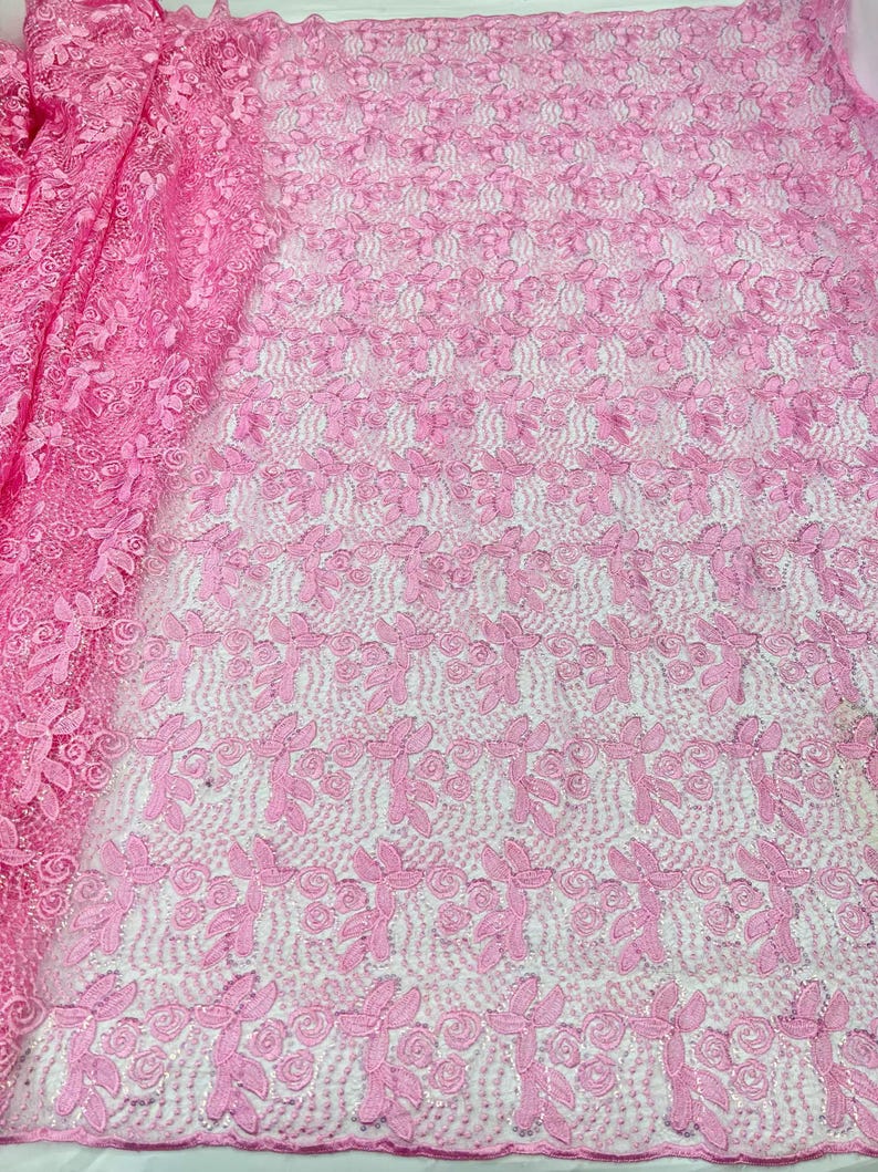 May include: A vibrant pink lace fabric featuring an intricate embroidered pattern of swirling floral motifs and leaves. The delicate mesh base is densely covered with raised thread work, creating a textured surface with a repeating botanical design.