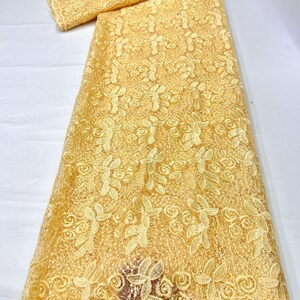 May include: A length of pale yellow lace fabric featuring an intricate embroidered pattern of swirling floral motifs and leaves. The textile has a delicate openwork mesh base with raised stitching that creates a textured, repeating design.