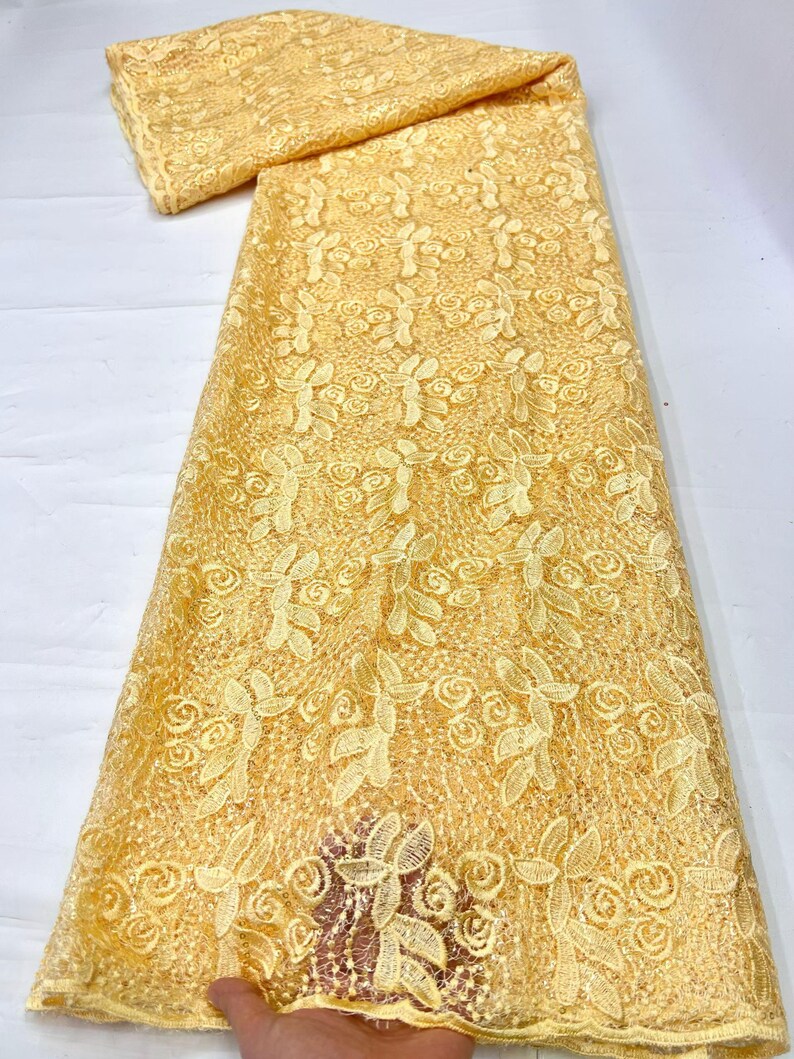 May include: A length of pale yellow lace fabric featuring an intricate embroidered pattern of swirling floral motifs and leaves. The textile has a delicate openwork mesh base with raised stitching that creates a textured, repeating design.