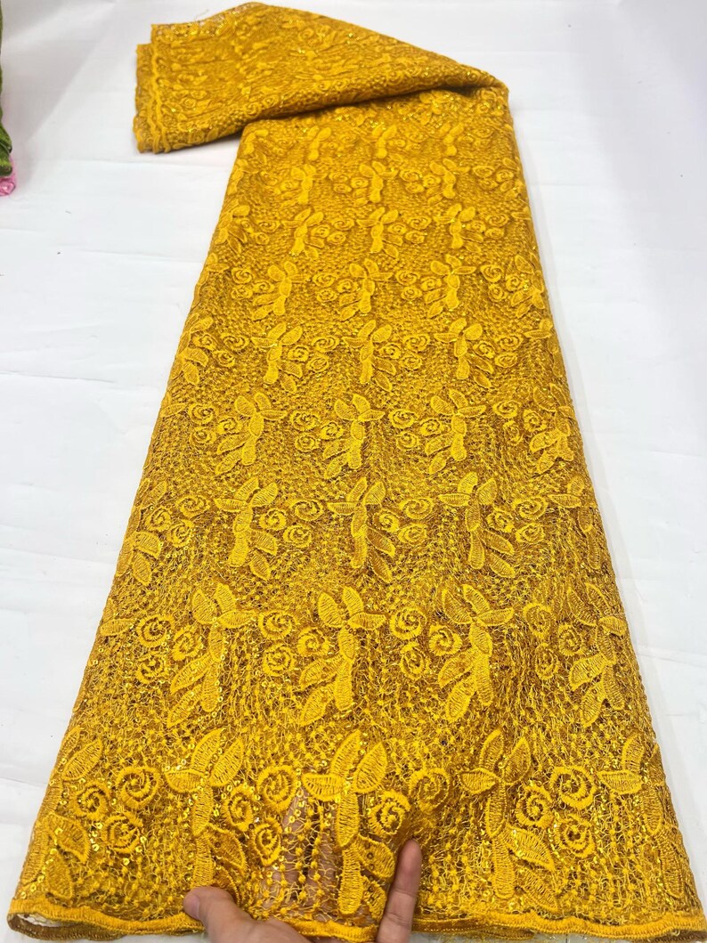 May include: A vibrant mustard yellow lace fabric featuring an intricate floral pattern with embroidered leaves and swirling rose-like motifs. The textile is embellished with small, shimmering sequins throughout the mesh base, creating a textured appearance.
