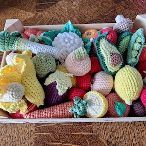 Peut inclure: Une cagette en bois remplie d'une variété de fruits et légumes au crochet, incluant un poireau, un chou-fleur, un avocat, des petits pois, une banane, une aubergine, un citron, une carotte et divers fruits ronds rouges et roses.