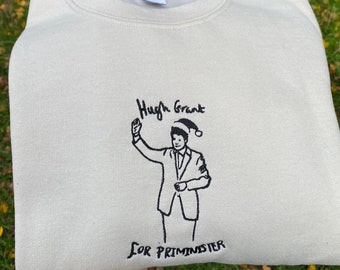 Op de afbeelding: Een crèmekleurig sweatshirt met een zwart geborduurd beeld van Hugh Grant met een kerstmanmuts en de tekst "Hugh Grant voor premier-minister".