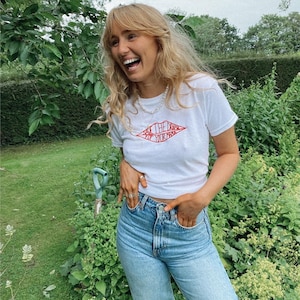 May include: A white t-shirt with the red text "Save the Drama for Your Mama". The t-shirt is worn with light blue jeans. The person is smiling in a garden setting.