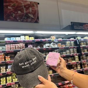 May include: A charcoal grey baseball cap with the embroidered phrase "hot girls just want aioli." The image also shows a package of roasted garlic aioli. The background is a supermarket with various food items on display.