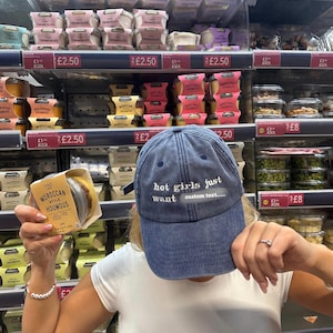 May include: A blue denim baseball cap with the text "hot girls just want custom text..." in white. The image also shows a person holding a package of Moroccan style houmous in a grocery store.
