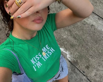 May include: A green t-shirt with the text "Kiss Me I'm Irish" in white lettering. The shirt is worn with a blue and white striped skirt and brown boots. The person is wearing a gold watch and several gold rings.