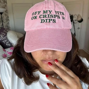 May include: A pale pink baseball cap featuring the embroidered phrase "OFF MY TITS ON CRISPS & DIPS" in dark green. The cap is worn by a person with dark hair and red nail polish.