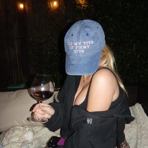 May include: A blue denim baseball cap with the embroidered text "OFF MY TITS ON PICKY BITS". A person is holding a glass of red wine. The person is wearing a black top and a leopard print skirt.