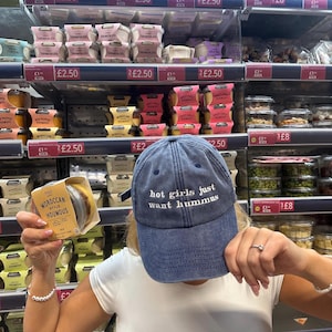 May include: A blue denim baseball cap with the white embroidered text "hot girls just want hummus". The image shows a person holding a package of Moroccan style hummus in a grocery store.