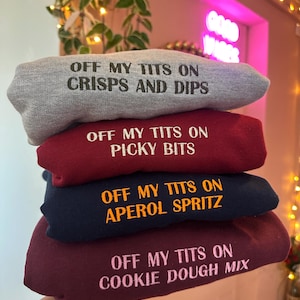 May include: Four sweatshirts in a stack, in grey, burgundy, navy, and maroon. Each has embroidered text: "OFF MY TITS ON CRISPS AND DIPS", "OFF MY TITS ON PICKY BITS", "OFF MY TITS ON APEROL SPRITZ", and "OFF MY TITS ON COOKIE DOUGH MIX".