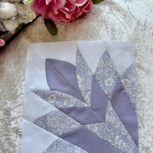 May include: A square quilt block featuring a stylized floral design. The design incorporates light purple and white fabric with a floral pattern. The block is made of various geometric shapes, creating a unique visual texture.