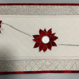 Könnte beinhalten: Gesteppter Tischläufer mit cremefarbenem Mittelteil, mit roten Poinsettien-Blumenapplikationen mit silbernen Zentren und Stielen. Die Ränder haben einen roten Rand und Zierleisten mit Muster, wodurch ein festliches Urlaubsdesign entsteht.