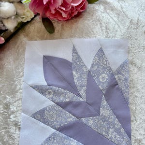 Peut inclure: Un bloc de tissu carré avec un motif floral. Le bloc présente un motif en forme d'éventail dans des tons de lavande et de blanc. Le motif intègre des pièces de tissu lavande uni et à motifs floraux.