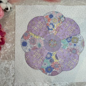 May include: A purple and white floral fabric flower with a white background. The flower is made of multiple fabric circles sewn together to create a flower shape.