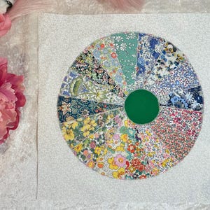 May include: A white fabric background with a colorful patchwork circle design. The circle is made of 16 different fabric pieces, each with a unique floral pattern. The center of the circle is a solid green circle.