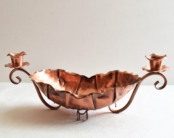 May include: A copper bowl with two candle holders on either side. The bowl is shaped like a flower and has a wavy edge. The candle holders are also shaped like flowers and have a swirl design.