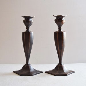 May include: A pair of antique bronze candle holders with a geometric design. The holders have a tall, slender base and a wide, flat top.