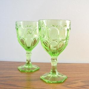May include: Two vintage green water goblets. The glasses have a decorative pattern of oval shapes and sit on a hexagonal base. The stem and base are a lighter shade of green than the bowl of the glass. The glasses are on a wooden surface.