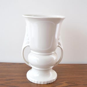 Könnte beinhalten: Eine weiße Keramikvase mit zwei gebogenen Griffen und einem ausgestellten oberen Rand. Die Vase hat einen bauchigen Mittelteil und einen Sockel. Die Oberfläche ist glatt und glänzend. Ein dekorativer Artikel für das Zuhause.