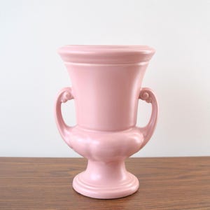 Könnte beinhalten: Eine rosa Keramikvase im Vintage-Stil mit einem Sockel und zwei geschwungenen Griffen. Die Vase hat eine breite, ausgestellte Oberseite und einen bauchigen Mittelteil. Die zarte rosa Farbe und das klassische Design machen sie zu einem dekorativen Stück.