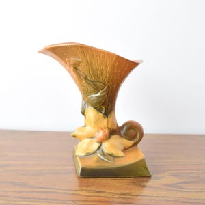 Pode incluir: Um vaso de cerâmica vintage em tons de castanho, laranja e verde. O vaso apresenta um design floral estilizado com um topo curvo e alargado e uma base retangular. A superfície texturizada adiciona interesse visual.