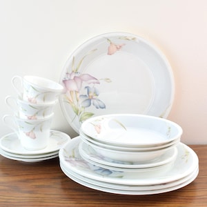 以下が含まれることがあります： 白い花柄の食器セット。お皿、ボウル、受け皿、ティーカップが含まれています。セットには、大きなサービングプレート、ディナープレート、ボウル、積み重ねられたティーカップとマッチするソーサーが含まれています。花柄のデザインにはピンクと青の花が含まれています。