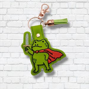 May include: A green frog keychain with a red cape and a sword. The frog is holding the sword and wearing a red cape. The keychain has a gold-colored clasp and a green tassel.
