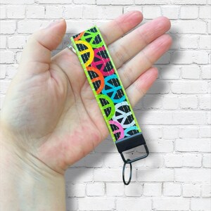 May include: A wrist lanyard with a black background and a repeating pattern of colorful peace signs in yellow, orange, pink, blue, and green. The lanyard has a black metal clip and ring.
