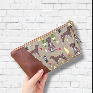 May include: A rectangular clutch with a brown leather panel and a tan fabric panel featuring a repeating pattern of brown dogs, wine bottles, and glasses. The clutch has a gold zipper and a brown tassel.