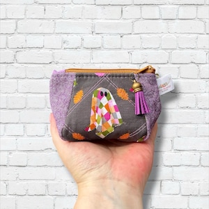 May include: A small fabric zipper pouch featuring a gray center panel with a colorful checkered ghost graphic and orange leaf accents. The pouch has purple side panels, a gold-toned zipper, and a small purple tassel attached to the zipper pull.