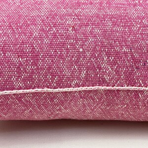 May include: A close-up of a pink pillow with a white stitched border. The pillow is made of a textured fabric with a subtle pattern.