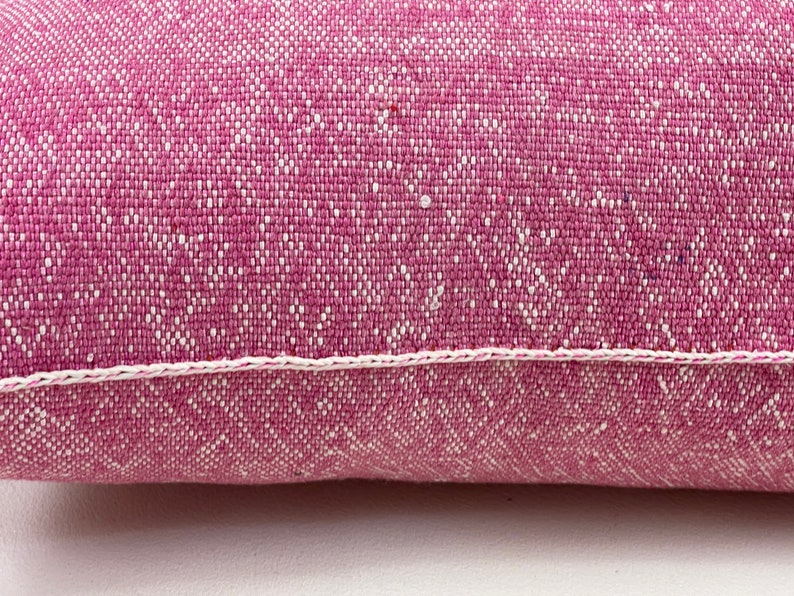May include: A close-up of a pink pillow with a white stitched border. The pillow is made of a textured fabric with a subtle pattern.