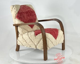 Puede incluir: Sillón tapizado con estructura de madera. La silla presenta una tela texturizada de color crema con detalles en forma de rombo rojo en el respaldo y el asiento. Los brazos y las patas de madera tienen un diseño curvo.