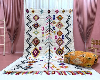 Peut inclure: Un tapis marocain blanc avec des motifs géométriques colorés et des franges. Le tapis a un motif en losange avec un motif d'arbre central. Un petit tapis orange avec un motif géométrique est au premier plan.