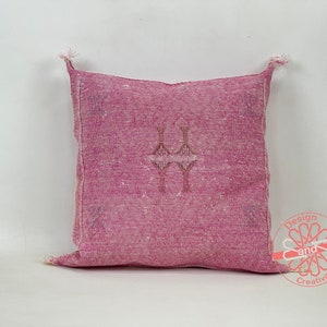 May include: A pink throw pillow with a traditional Moroccan design. The pillow has a woven texture and a fringe border.