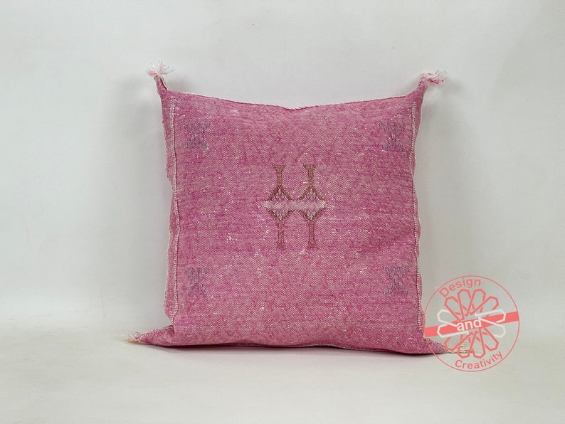 May include: A pink throw pillow with a traditional Moroccan design. The pillow has a woven texture and a fringe border.