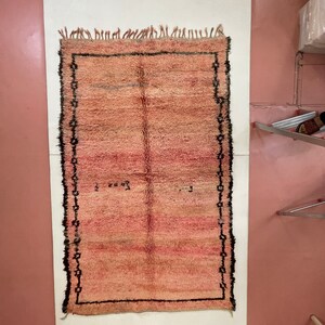 Op de afbeelding: Een handgeweven roze en beige tapijt met een zwarte rand. Het tapijt heeft aan alle kanten franjes.