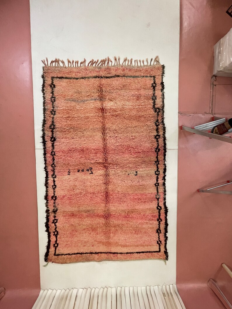 Op de afbeelding: Een handgeweven roze en beige tapijt met een zwarte rand. Het tapijt heeft aan alle kanten franjes.