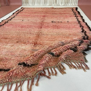 Op de afbeelding: Een roze en zwart Marokkaans tapijt met franjes. Het tapijt is dubbelgevouwen en heeft een zachte, pluche textuur.