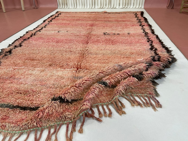 Op de afbeelding: Een roze en zwart Marokkaans tapijt met franjes. Het tapijt is dubbelgevouwen en heeft een zachte, pluche textuur.