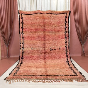 Op de afbeelding: Een groot, handgeweven tapijt in roze en beige tinten. Het tapijt heeft een zwarte rand met een geometrisch patroon en een franje aan de onderkant.