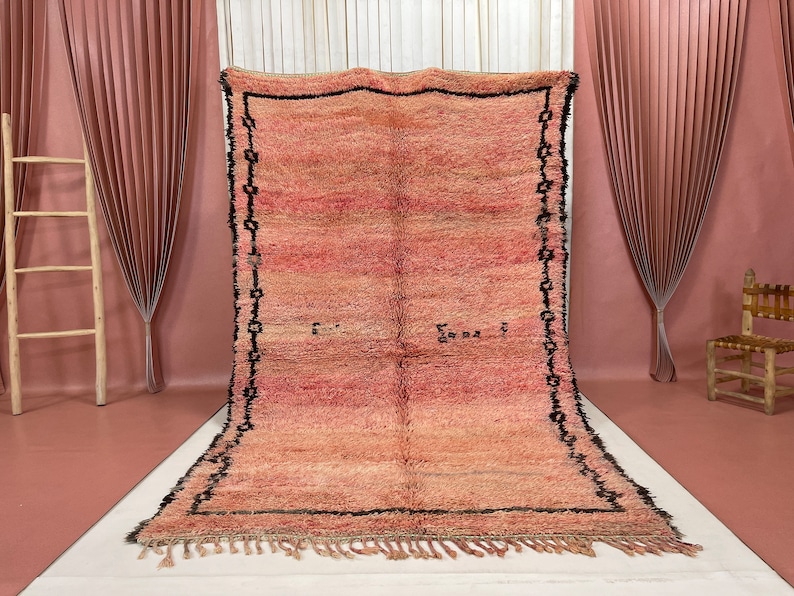 Op de afbeelding: Een groot, handgeweven tapijt in roze en beige tinten. Het tapijt heeft een zwarte rand met een geometrisch patroon en een franje aan de onderkant.