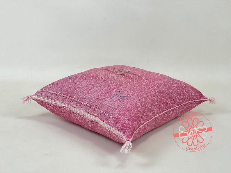May include: A pink and white patterned floor cushion with a fringe detail. The cushion is made of a soft, woven fabric and has a subtle sheen.