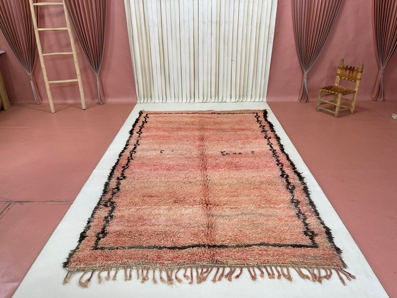 Op de afbeelding: Een roze en cr&egrave;me Marokkaans tapijt met een zwarte rand en franjes. Het tapijt heeft een verweerde look en is gemaakt van wol.