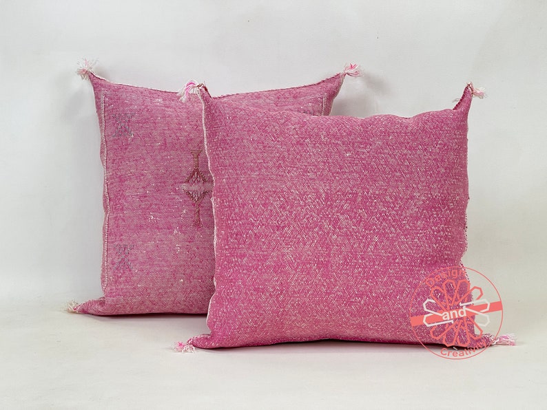 May include: Two pink throw pillows with a subtle woven pattern and fringe detailing. The pillows are made of a soft, textured fabric.