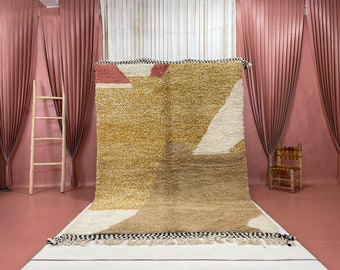 May include: A large, abstract area rug with geometric shapes in shades of beige, brown, white, and pink. The rug has a fringe border and is displayed on a pink floor.