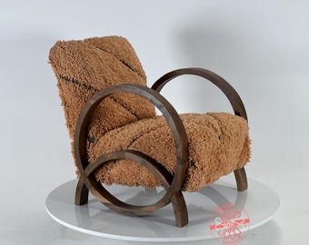Puede incluir: Un sillón marrón con un marco de madera curvado y una tapicería de tela marrón esponjosa. El sillón está sobre un pedestal blanco.